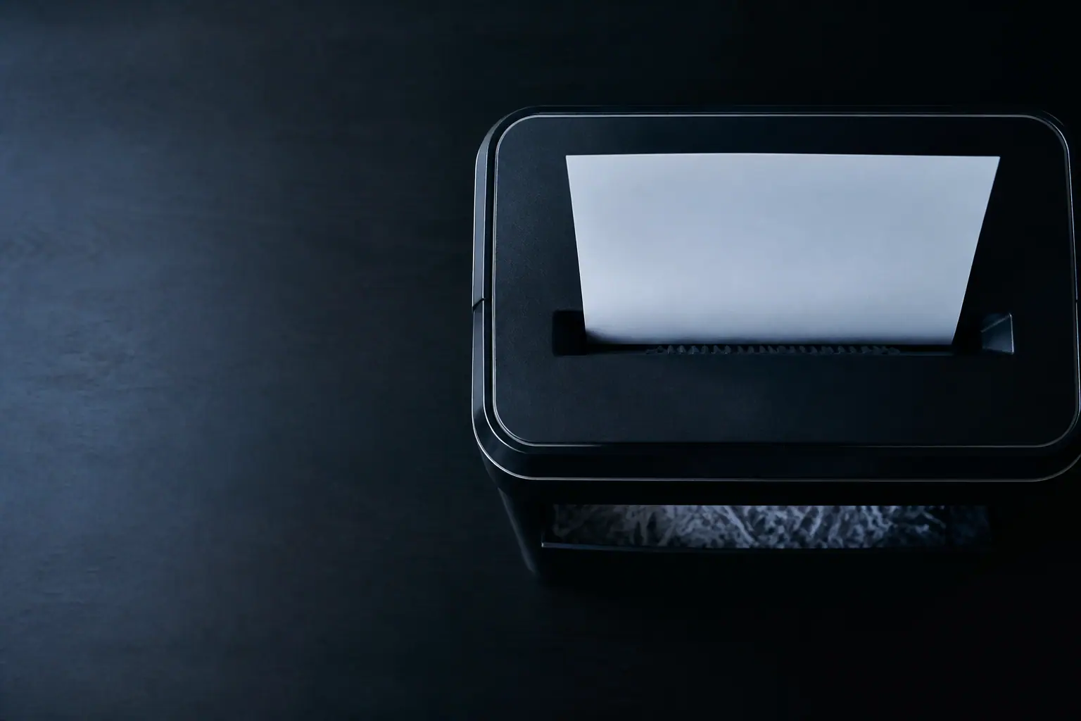 An overhead view of a blank sheet of paper being drawn into an office shredder on a dark desk.
