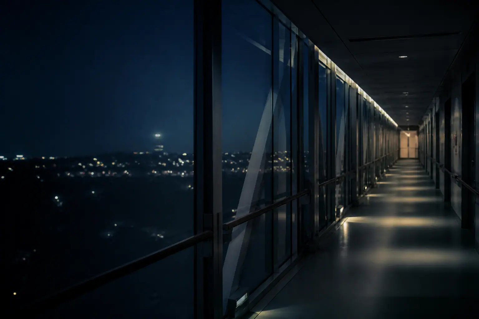 A long hospital corridor receding into blue evening light, seen through glass.