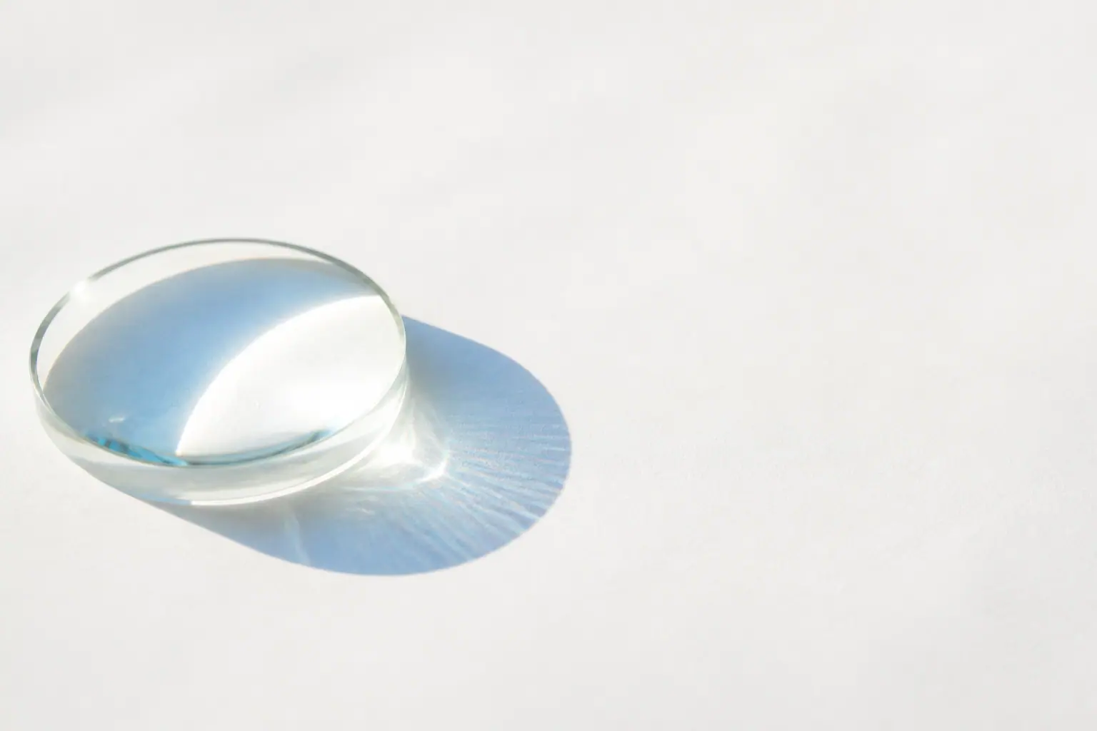 A clear glass lens resting on white paper, casting a soft pale-blue shadow in daylight.